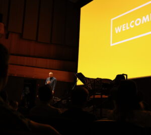 will guidara speaks on stage at welcome conference, yellow welcome conference logo projected behind him