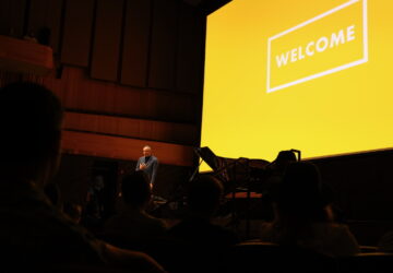 will guidara speaks on stage at welcome conference, yellow welcome conference logo projected behind him